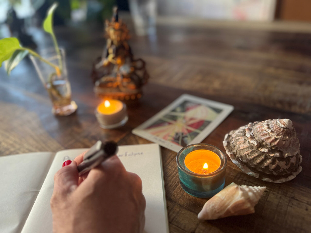 Journal with a journaling hand, full moon lunar eclipse journaling, a plant, shells that symbolize the water of Pisces, ritual objects, a goddess statue and the Tarot card The Hermit that represents the Virgo archetype - connecting myth and meaning.